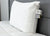 A white Manchester Mills Envirosleep Dream Support Pillow with fabric tags stands upright against a gray upholstered headboard on a white quilted mattress, creating a clean, organized look in a modern bedroom.
