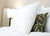 A Pillowtex Premium Polyester Pillow | Extra Firm by Pillowtex with a visible care tag leans against a green pillow with a black floral pattern on a white bed. Behind them, a lit table lamp and gold-framed mirror stand against a light wall.