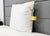 The JS Fiber Super Gold Choice Pillow | Heavyweight by JS Fiber, a white square pillow with yellow trim and tag for side sleepers, stands upright against a gray headboard on a neatly made bed with a white comforter in a minimal setting.