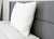 A Pillow Factory Comforel Pillow, featured at Sheraton Hotels, with a textured white cover and care label, sits upright against a gray upholstered headboard on a neatly made bed with a white quilted mattress cover in a clean, minimal setting.