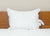 The Pacific Coast® Natural Feather Pillow by Pacific Coast Feather Company, featuring subtle floral patterns, sits upright against a wooden headboard on a white quilted mattress. The pillow has tags on the right side and a beige wall in the background.