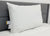 The Envirosleep Gold Plus Pillow by Manchester Mills, featuring firm polyester fiber fill and attached care tags, stands upright on a neatly made bed with a white comforter and modern gray upholstered headboard.