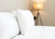 A neatly made bed with white bedding features the Keeco Best Western® Everest Pillow, a soft polyester pillow, near a beige wall. A tripod floor lamp with a woven shade glows warmly in the background, creating an inviting atmosphere.