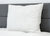 A large, square Pillow Factory Comforel Pillow, featured at Sheraton Hotels, with subtle horizontal stripes leans against a gray headboard on a white quilted mattress; the background wall is plain white.