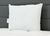 The JS Fiber Hollofil II Pillow | Medium by JS Fiber, a square white hypoallergenic pillow with a subtle fabric texture, sits upright against a dark gray padded headboard on a white quilted bed. A small fabric tag is visible on its left side.