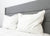 Close-up of a neatly made bed featuring the Pillowtex Hotel Feather and Down Pillow Set (2 pillows) by Pillowtex, with fluffy white pillows, white bedding, a light gray upholstered headboard, and a minimalist background.