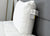 A white Envirosleep Dream Support Pillow by Manchester Mills with medium support and two labels rests against a gray headboard on a neatly made bed with a white comforter, with part of a mirror and white wall visible in the background.