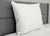 The Down Etc. Fairfax Pillow—featured at St. Julien Hotel & Spa<sup>®</sup>—for side and back sleepers, is propped upright against a gray headboard on a neatly made bed with a white comforter; its care tag is visible. A light gray wall forms the background.