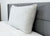 A firm Envirosleep Gold Plus Pillow by Manchester Mills, featuring polyester fiber fill, rests upright against a modern gray headboard. The bed is topped with a white quilted comforter and set before a light wall for a clean, minimal look.