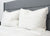 Two Pillowtex Triple Core White Duck Down & Feather Pillows with medium-soft support rest side by side against a gray headboard. The bed features a white quilted comforter, offering a clean, minimal look ideal for side and back sleepers. Pillow tag visible.