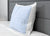 A single Pillowtex pillow with the Pillowtex Cooling Gel Pillow Protector (light blue, grid-patterned) rests upright against a gray upholstered headboard on a white mattress, creating a minimalist and comfortable bedroom setting.