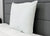 A medium white JS Fiber Hollofil II Pillow from JS Fiber, featuring a smooth texture, stands upright against a dark gray headboard on a quilted bedspread. This hypoallergenic pillow looks plush and clean in a modern bedroom.