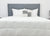 A bed with a gray upholstered headboard features a white comforter, three large white pillows, two striped throw pillows, and is topped with the Pillowtex Hotel Blanket by Pillowtex for added comfort. A plant decorates the right nightstand.