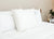 A neatly made bed features two large white pillows and a Best Western® Aurora Pillow | Medium Polyester Pillow by Keeco. A small potted plant sits on a wooden nightstand near a beige wall.