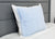 A Pillowtex Cooling Gel Pillow Protector covers a pillow with a light blue, grid-patterned case and white border, resting upright against a gray upholstered headboard on a white comforter for a clean, modern bedroom look.