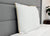 The JS Fiber Super Gold Choice Pillow | Heavyweight by JS Fiber, a square white pillow with yellow trim, is ideal for side sleepers and rests against a gray upholstered headboard on a neatly made bed with a wooden nightstand and potted plant nearby.