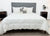 A neatly made bed with a gray upholstered headboard and white comforter features three white pillows, two striped accent pillows, and the Pillowtex Hotel Blanket by Pillowtex. Modern nightstands hold clocks and potted plants to complete the minimalist room.
