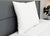 A Pillow Factory Comforel Pillow, featured at Sheraton Hotels, is propped against a gray headboard on a neatly made bed with a white comforter. A small potted plant sits on the white bedside table in the background.