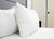 A close-up of two Downlite Primaloft Down Alternative Pillows stacked on a neatly made bed with a white quilted mattress cover, light grey padded headboard, and a white closet door in the background. The scene looks clean and tidy.