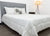 A neatly made bed with a gray upholstered headboard, white bedding, and the Pillowtex Hotel Blanket by Pillowtex. Two white pillows and a black-and-white accent pillow rest beneath a standing lamp against crisp white walls and wood floors.