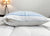 A close-up of a Pillowtex Cooling Gel Pillow Protector in white with a blue quilted top, partially unzipped to reveal the filling. A lamp and nightstand are seen in the background against a neutral wall.