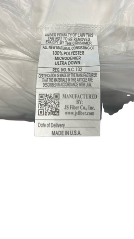 A close-up view of a bedding label showing material composition, manufacturer details, and QR code, indicating it's made of 100% polyester with Ultra Down Pillow filling, by JS Fiber "Ultra Down" Polyester Pillow | Firm.