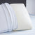 A white Carpenter Comfort Tech Bed Pillow Featuring Tranquility Foam with small ventilation holes stands upright on a bed, partially covered by a quilted white blanket with blue trim, atop a light blue and white striped mattress and dark gray headboard.