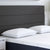 A close-up of a modern bed with a dark gray upholstered headboard, two Carpenter Comfort Tech Bed Pillows featuring Tranquility Foam, and a subtly patterned white mattress. A small white nightstand is partially visible on the left.