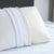 A close-up of the Carpenter Comfort Tech Bed Pillow Featuring Tranquility Foam, designed for side sleepers with ventilation holes, partially covered by a white pillowcase and resting on a light blue striped mattress against a gray headboard.