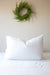 A white, fluffy Pillow Factory® 75/25 Duck Feather/Down Pillow, perfect for back and side sleepers, is centered on a neatly made bed with white sheets. Above the pillow, a green fern wreath hangs on a beige wall, adding a touch of nature to the otherwise minimalistic and clean space. The scene conveys a sense of calm and simplicity.