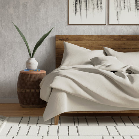 A cozy bedroom corner with an unmade bed, featuring a Delilah Home Organic Cotton Duvet Cover made of hypoallergenic fabric and a wooden headboard. A woven basket nightstand holds a vase with a plant.
