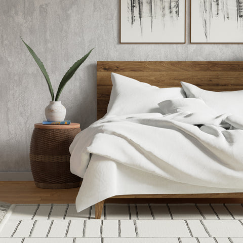 A cozy, contemporary bedroom with a wooden bed frame, white 300-count bed sheets, and a woven bedside table adorned with a plant in a white vase, complemented by neutral-toned wall art and a Delilah Home Organic Cotton Duvet Cover.