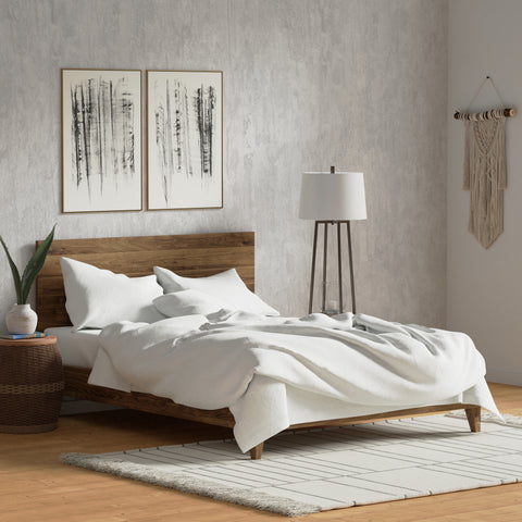 A minimalist bedroom featuring a wooden bed with white Delilah Home Organic Cotton Duvet Cover bedding, abstract art on the concrete wall, a simple lamp on a side table, a macrame wall hanging, and a textured rug.
