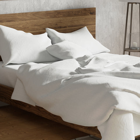 An inviting, unmade bed with crumpled white Delilah Home Organic Cotton Sheet Set on a wooden bed frame, suggesting a recent departure or a lazy morning in a room with a simple, minimalist aesthetic. The bed is
