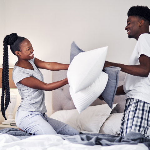 A man and woman are playfully tossing Encompass Group 50/50 Gray Duck Feather and Down Pillows on the bed, finding comfort in each other's support.