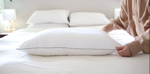 A person in a beige hoodie adjusts a Carpenter Indulgence® Synthetic Down Pillow on a neatly made bed with white linens. The background shows a bedside table with a lamp, enhancing a cozy atmosphere.