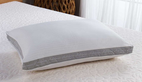 A white, rectangular Carpenter Indulgence® Synthetic Down Pillow | Side Sleeper with textured stripes on its surface, prominently displaying a gray embroidered label with elegant script around the edge, placed on a textured white bedspread against a softly lit background.