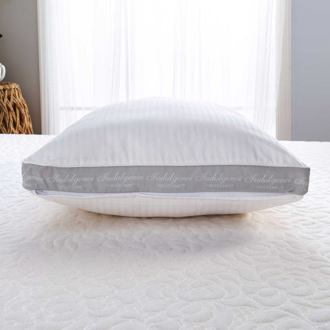 A plush white Indulgence® Synthetic Down Pillow | Side Sleeper by Carpenter with "indulgence" repeated on its trim sits on a textured white bedspread. Behind the pillow, a sheer curtain softly diffuses daylight, creating a serene atmosphere.