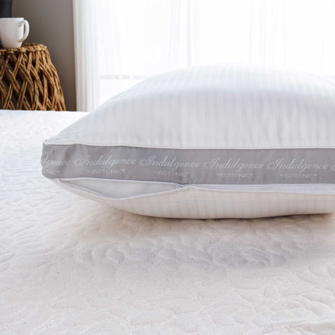 A close-up image of a white striped, machine-washable Carpenter Indulgence® Synthetic Down Pillow with the word "indulgence" repeatedly printed on its fabric. The pillow rests on a textured white bedspread and is illuminated.