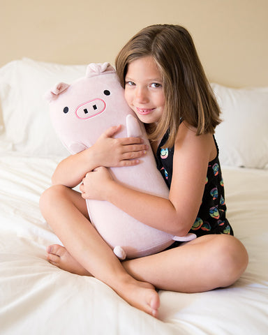 A girl sitting on a bed with a Memory Foam Pig Themed Pillow, Wilbur The Pig by Pillowtex, a perfect gift for children.