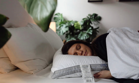 A woman sleeps peacefully under a white, machine-washable comforter in a cozy bedroom with light beige sheets. Lush green plants add freshness to the room’s tranquil ambiance, while she cuddles up with her Carpenter Indulgence® Synthetic Down Pillow designed for side sleepers.