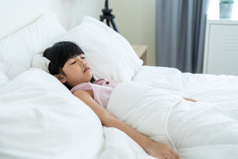A young Asian girl is peacefully asleep on a white bed with an Encompass Group 50/50 Gray Goose Feather and Down Pillow.