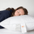 A woman with long hair peacefully rests her head on a white Holiday Inn<sup>®</sup> Soft Support Polyester Pillow by Hollander, dressed in a dark blue top. The pillow, brimming with polyester fill, boasts a tag displaying the IHG logo along with care instructions. A plain white wall in the background accentuates the minimalist atmosphere, completing her serene and relaxed appearance.