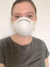 A person is facing the camera, wearing a white *pillowsdotcom* Final Sale: NIOSH N95 Respirator | Pack of 20 Masks that covers the nose and mouth. They have light skin, light-colored eyes, and brown hair pulled back. They are wearing a gray t-shirt and the background appears to be a plain, white wall.